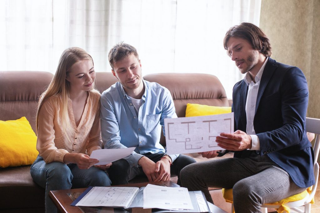 Professional property manager showing house plan to young couple indoors